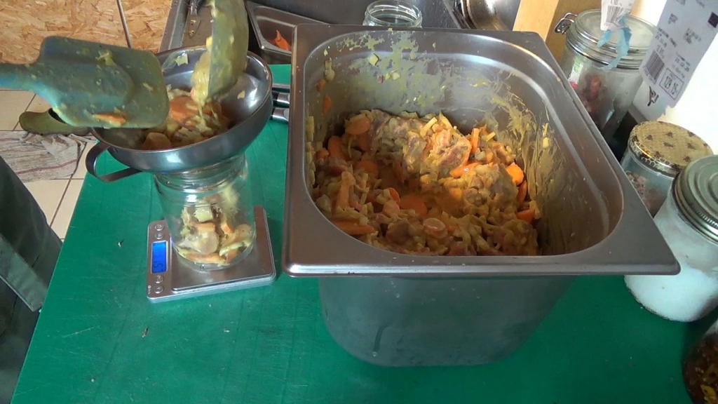 Mise en bocal d'un porc coco curry aux carottes avec un entonnoir, le bocal posé sur la balance